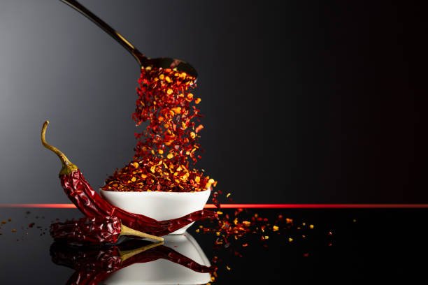 Dry chili pepper flakes in a small ceramic bowl on a black reflective background. Red pepper flakes pour in white bowl. Copy space.